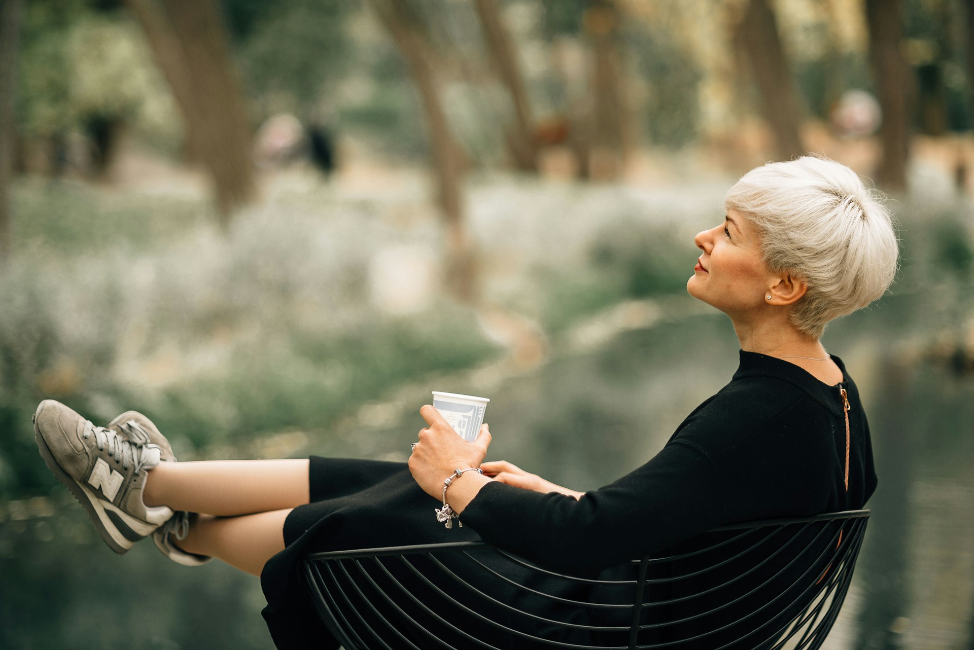 Woman with short platinum hair and a black dress relaxing in a metal chair, holding a cup, legs stretched out and gazing upward in a leafy park by the water.